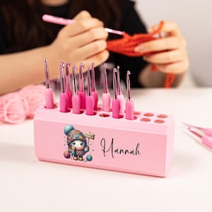 May include: A pink crochet hook holder with a cartoon character and the name "Hannah" printed on the front. The holder contains multiple pink crochet hooks. A person is crocheting with an orange yarn in the background.