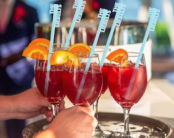 May include: Four wine glasses filled with a red beverage, garnished with orange slices and light blue straws. The straws have the text "THE LIEBELTS" printed on them. The glasses are held on a silver tray.