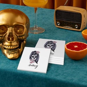 Peut inclure: Un crâne doré, des dessous de verre avec un motif de tête de mort, une radio vintage et un demi-pamplemousse sont posés sur une surface turquoise. Les dessous de verre présentent une tête de mort avec les noms "Aubrey" et "Lia". Un verre de liquide orange est également présent.