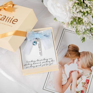 May include: A beige gift box with a gold ribbon and the name "Carrie" printed on it. Inside, a card with a blue charm and the text "October 25, 2023". The card also has the text "Something old something new..."