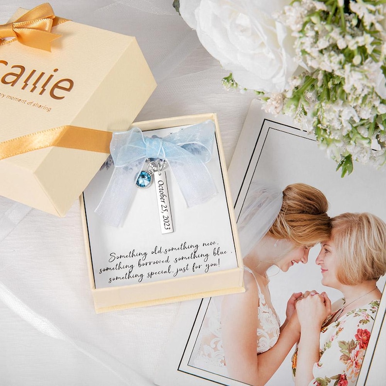 May include: A beige gift box with a gold ribbon and the name "Carrie" printed on it. Inside, a card with a blue charm and the text "October 25, 2023". The card also has the text "Something old something new..."