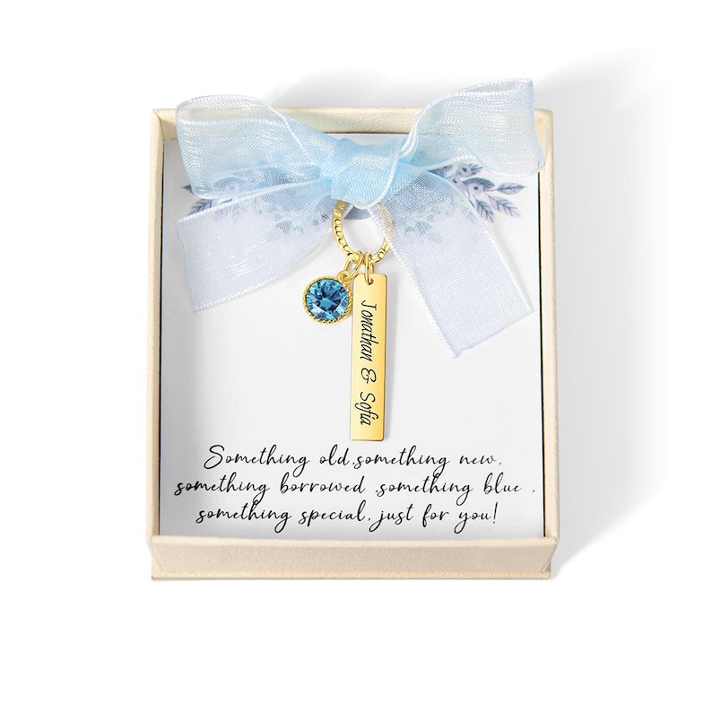 May include: A gift box with a light blue ribbon and a gold-colored charm. The charm has a blue gemstone and a rectangular tag with the names "Jonathan & Sofia". The box contains the text "Something old something new..."