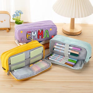 May include: Three rectangular pencil cases in purple, yellow, and light blue. The purple case features the name EMMA in colorful letters with star and apple accents. The yellow and light blue cases are open, revealing internal mesh pockets and elastic loops holding various markers and sticky notes.