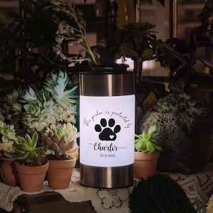 Puede incluir: Lámpara de jardín cilíndrica de acero inoxidable con un panel blanco brillante. El panel presenta una huella de pata negra, el texto "This garden is protected by", el nombre "Charlir" con un corazón y los años 2015-2025.