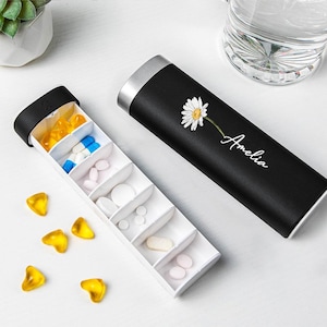 Può includere: Un portapillole nero con parte superiore argentata e un design a margherita bianca, personalizzato con il nome "Amelia". L'organizzatore aperto rivela più scomparti pieni di varie pillole e capsule. Alcune pillole gialle a forma di cuore sono sparse.