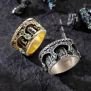 May include: Two ornate rings, one gold-toned and one silver-toned, are displayed. The gold ring features black detailing, teal gemstones, and the letter "L". The silver ring has green gemstones and the letter "A". Both rings have intricate designs.