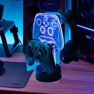 Peut inclure: Un ensemble d'accessoires de jeu avec un support en acrylique transparent en forme de manette, éclairé par des LED bleues. Une manette noire repose sur le support, avec un casque suspendu. L'ensemble est sur un bureau en bois, créant une esthétique technologique.