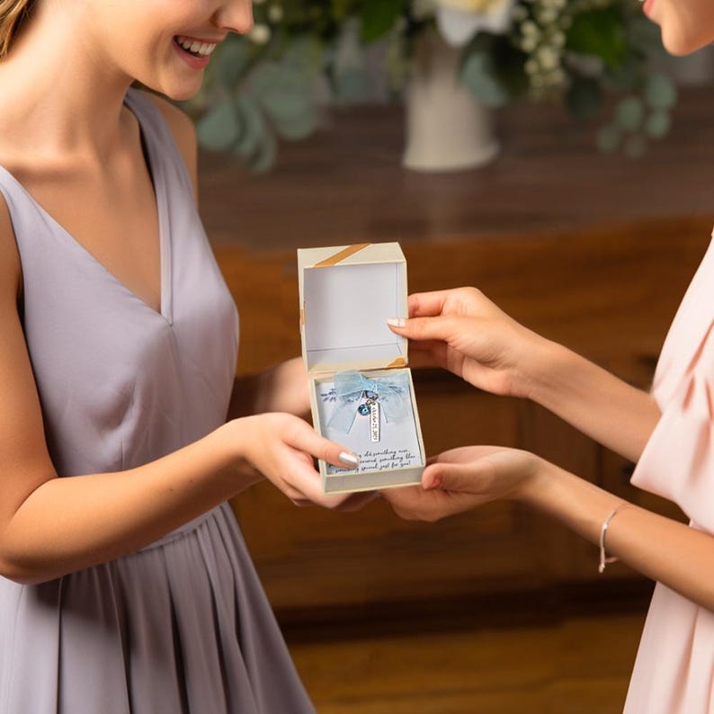 May include: A small, open gift box with a gold ribbon on top is held by two people. Inside, a necklace with a blue charm and the name "Nick" is visible, resting on a blue ribbon. The box also contains a small card with text.