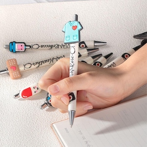 Puede incluir: Colección de bolígrafos con adornos médicos, incluyendo una bata azul con un corazón rojo, una venda, una pastilla y un dispositivo médico. Los bolígrafos tienen los nombres "Rebecca" y "Heather" impresos.