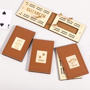 May include: A wooden cribbage board engraved with the name "ELIZABETH" and the text "EST. 2021". Four brown leather card holders are also present, each with a wooden insert. The inserts feature the text "VON LIFE Cleake", paw prints with the name "Richard", and the name "Garcia".