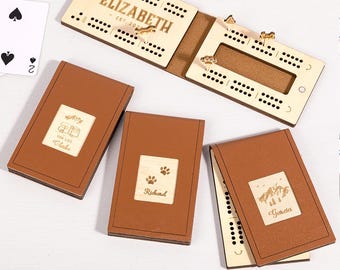 May include: A wooden cribbage board with the name "ELIZABETH" and the text "EST. 2021" engraved on it. There are also four brown leather-bound card holders with personalized wooden inserts. One insert has the text "VON LIFE Cleake", another has paw prints and the name "Richard", and the third has the name "Garcia".