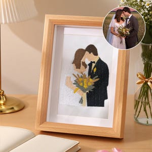 Puede incluir: Un marco de fotos de madera clara que contiene un retrato bordado de una pareja con traje de boda. El bordado representa a una persona con vestido blanco y a otra con traje negro sosteniendo un ramo de flores amarillas y verdes.