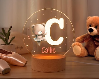 May include: A personalized night light with a wooden base. The clear acrylic light features a large white letter "C", a cartoon bear ballerina, and the name "Callie". The light is surrounded by stars and hearts.