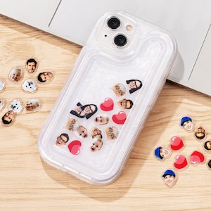 May include: Clear phone case with floating face cutouts and red heart shapes. The case is on a wooden surface with additional face cutouts and hearts scattered around. The phone case has a dual-camera system.