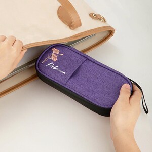 May include: A purple pencil case with the name "Rebecca" in cursive and a floral design. The case has a black base and a small black handle. The case is being held near a beige tote bag.