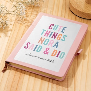 Puede incluir: Un cuaderno rosa con el texto "CUTE THINGS NORA SAID & DID when she was little" en letras coloridas. El cuaderno tiene una cinta de marcador y un cierre de correa rosa. El cuaderno está sobre una superficie de madera.