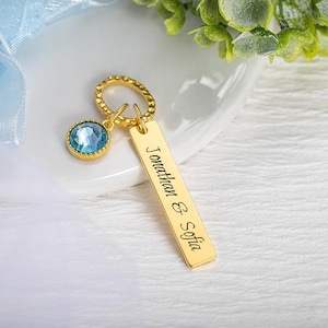 May include: A gold-colored keychain with a rectangular tag engraved with the names "Jonathan & Sofia". It features a round, light blue gemstone charm and a textured ring. The keychain is displayed on a white surface with greenery in the background.