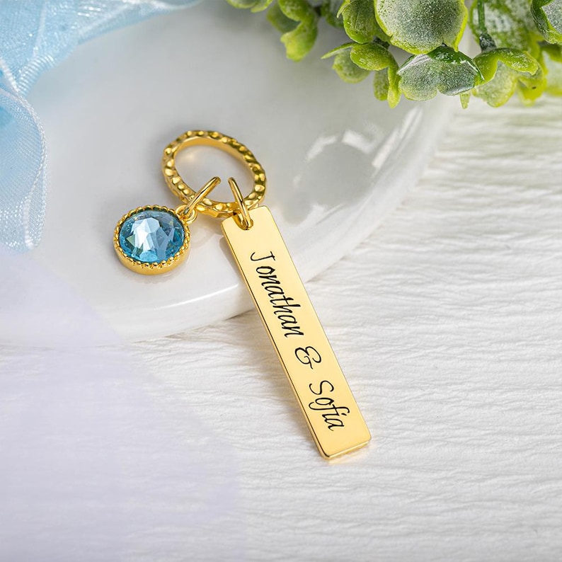 May include: A gold-colored keychain with a rectangular tag engraved with the names "Jonathan & Sofia". It features a round, light blue gemstone charm and a textured ring. The keychain is displayed on a white surface with greenery in the background.