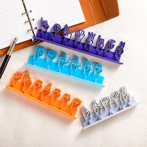 Könnte beinhalten: Eine Sammlung farbenfroher ASL (American Sign Language) Namensschilder. Die Zeichen sind in Lila, Blau, Orange und Grau gehalten. Die Namen werden mit Handgesten buchstabiert. Im Hintergrund befinden sich ein Notizbuch und ein Stift.