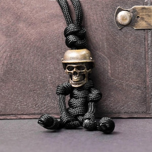 May include: A bronze-colored skull pendant wearing a helmet with the word "IMAN" on it, attached to a black woven cord. The skull is detailed with visible teeth and eye sockets. The cord is knotted at the top and forms the body and limbs of the skull.