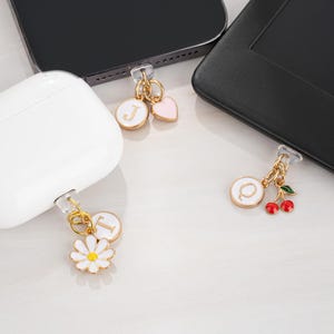 Może przedstawiać: Zbliżenie na trzy zawieszki do telefonu ze złotymi łańcuszkami. Jedna zawieszka ma białe kółko z literą "J" i różowym sercem. Druga ma białą stokrotkę i literę "T". Trzecia ma białe kółko z literą "Q" i czerwonymi wiśniami.