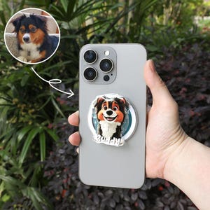 May include: A silver smartphone with a pop-up phone grip featuring a cartoon dog with the name "Buddy". The dog has brown, black, and white fur. An image of the dog is in a circle above the phone.