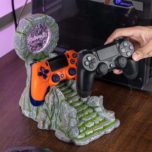 May include: A gray stone-like gaming controller stand with a circular portal featuring the text "Shadowfall". An orange and blue controller rests on the stand, with a black controller held in a hand. The stand has a mossy, rocky design.