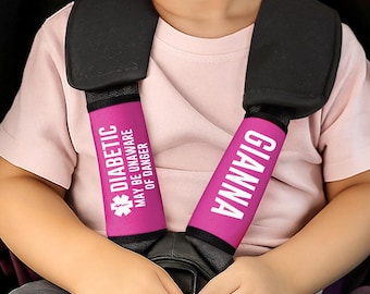 May include: Pink seat belt covers with white text. One cover reads "DIABETIC MAY BE UNAWARE OF DANGER" with a medical symbol. The other cover says "GIANNA". The covers are on a child's car seat.