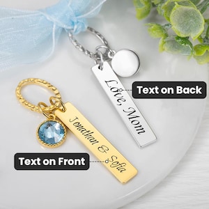 May include: Two personalized keychains. One gold-colored keychain with a blue gemstone and the names "Jonathan & Sofia" engraved. The other is silver-colored with "Love Mom" engraved. Both have a rectangular tag design.