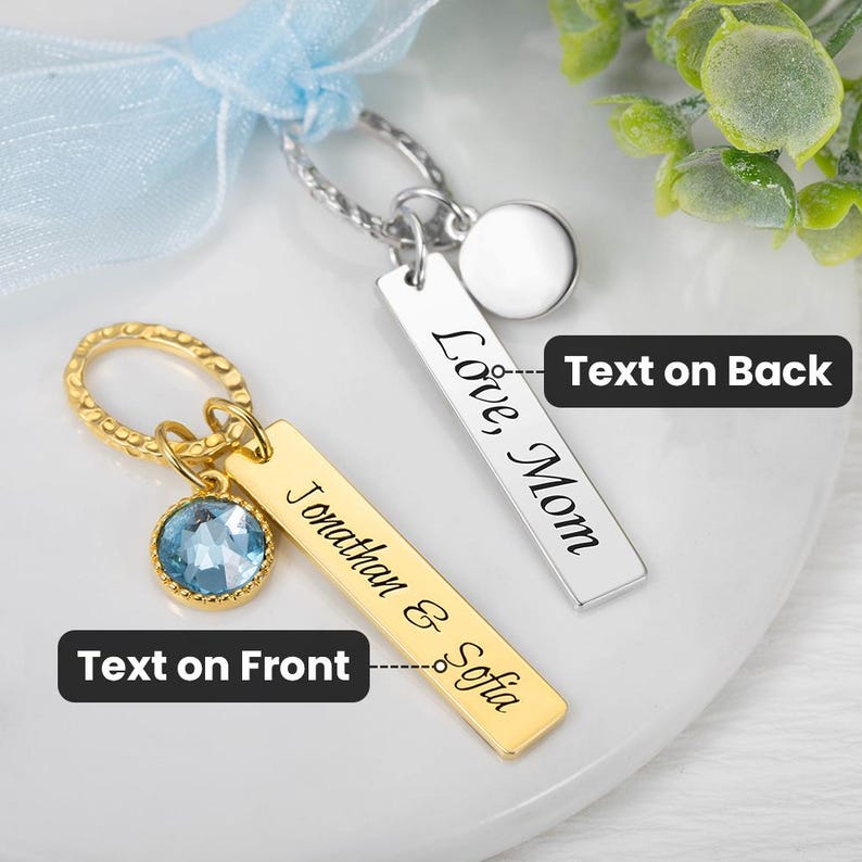 May include: Two personalized keychains. One gold-colored keychain with a blue gemstone and the names "Jonathan & Sofia" engraved. The other is silver-colored with "Love Mom" engraved. Both have a rectangular tag design.