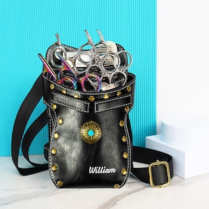 Op de afbeelding: Zwarte leren kappersgereedschapsholster met goudkleurige klinknagels en een decoratief turquoise middenstuk. De holster bevat meerdere scharen en een kam. De naam "William" staat op de voorkant. Inclusief zwarte riem en gouden gesp.