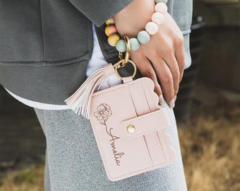 May include: A light pink cardholder with a floral design and the name "Amelia" is attached to a gold keyring and tassel. A beaded bracelet with pastel colors is worn on the wrist. The cardholder has a scalloped edge and a snap closure.