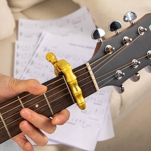 Op de afbeelding: Een goudkleurige gitaarcapo met een uniek ontwerp is bevestigd op de toets van een akoestische gitaar. De capo heeft de naam "Brian" gegraveerd. Bladmuziek is zichtbaar op de achtergrond.