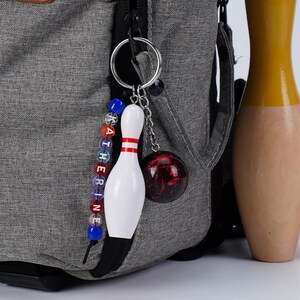 Puede incluir: Un llavero con temática de bolos con un pin blanco y un dije de bola de bolos roja y negra. El llavero está sujeto a una mochila gris. Una cadena de cuentas deletrea el nombre "KATHERINE".