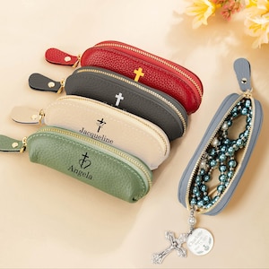 May include: A collection of zippered leather rosary cases in red, gray, cream, and green, each with a cross emblem. One case is open, revealing a blue rosary. A silver cross pendant with a circular charm is attached.