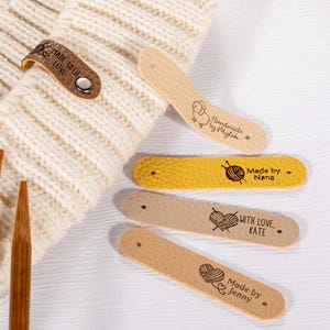 May include: A collection of leather tags with various text and designs. One tag reads "MADE WITH LOVE" and is attached to a cream-colored knitted item. Other tags say "Handmade by Mylah", "Made by Nana", "WITH LOVE, KATE", and "Made by Jenny".