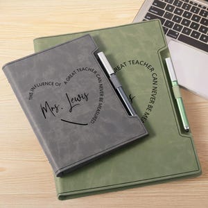 Op de afbeelding: Twee gepersonaliseerde cadeaus voor leraren: een grijze en een groene portfolio van imitatieleer. De grijze portfolio heeft de tekst "De invloed van een geweldige leraar kan nooit worden gemeten" en "Mrs. Lewis". Twee pennen zijn inbegrepen.