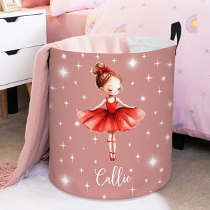 May include: A pink storage bin featuring a ballerina illustration in a red dress and a matching hair bow. The name "Callie" is written in cursive below the ballerina. White starbursts are scattered across the pink background.