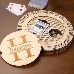Op de afbeelding: Een houten cribbagebord met een gepersonaliseerd deksel met de naam "Johnston" en de initiaal "H". Het bord bevat een pak speelkaarten en metalen pinnen. Een gedeeltelijk kaartspel is zichtbaar.