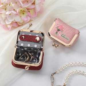 May include: Two small, textured leather pouches with gold-tone metal frames and kiss-lock closures. One pouch is open, revealing a grey interior with white polka dots and two metal rings. The other is closed, featuring a pink floral print and the name Glaria.