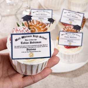 Pode incluir: Um cupcake com cobertura branca, coberto com um diploma e um chapéu de formatura. O diploma diz "Walt Whitman High School" e "Tobias Dadisman". Outros cupcakes com diplomas estão no fundo.