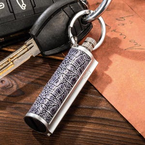 May include: A silver and grey keychain lighter with a textured, crocodile-style pattern. The lighter is attached to a key ring and has the word "Nelly" printed on the side. The lighter is next to a car key.