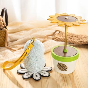 May include: A light blue yarn ball with a yellow wrist strap that reads "Oallie's crochet" sits on a white and black flower-shaped coaster. A sunflower-shaped yarn holder and a basket are also in the image. The yarn holder has a gold stem and a brown and yellow flower.