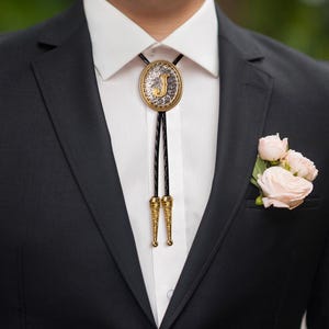 May include: A gold and silver bolo tie featuring the letter "J", worn with a white shirt and black suit. The tie has a braided black cord and decorative gold tips. A small bouquet of pink roses is in the suit pocket.