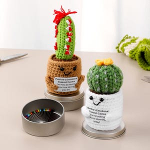 May include: Two handmade crochet cactus ornaments, one green with red accents and the other green with a yellow flower. Each cactus is in a pot with a smiling face and a supportive message. A small tin with colourful pins is in the foreground.