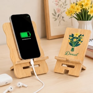May include: Two light-colored bamboo phone stands with a wavy edge design. One stand holds a black smartphone displaying a green battery icon and the text Current battery level 74%. The other stand features a printed bouquet of yellow daffodils and the name Daniel in green cursive script.