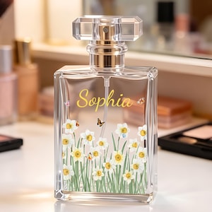Op de afbeelding: Een rechthoekige, heldere glazen parfumfles met een transparante dop en een zilverkleurige verstuiver. De voorzijde toont de naam Sophia in geel schrift, omringd door een print van witte narcissen met gele harten en kleine vlinders.