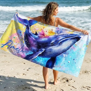 Puede incluir: Una toalla de playa rectangular con un diseño de ballena azul con un patrón de galaxia. La toalla tiene el nombre "Amelia" en escritura cursiva. La toalla se sostiene en una playa de arena.
