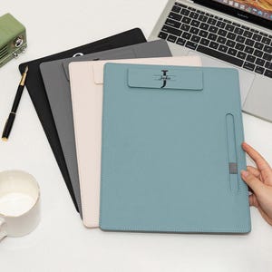 May include: A stack of four leather clipboards in black, gray, blush pink, and teal. The teal clipboard has the name "Jackie" printed on it. A pen, a mug, and a laptop are also visible.