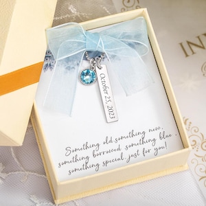 May include: A light blue pendant necklace with a round, faceted gemstone and a rectangular tag engraved with "October 25, 2023." The necklace is presented in a cream-colored gift box, tied with a light blue ribbon. Text on the card reads "Something old, something new..."
